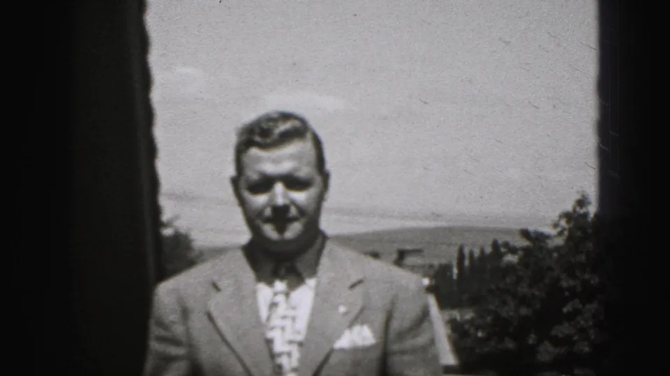 The man faces the camera in a light suit and patterned tie, with trees, rooftops, sky, and distant hills behind him.