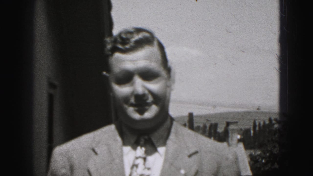 Suited man walks closer to the camera with a slight smile outdoors.
