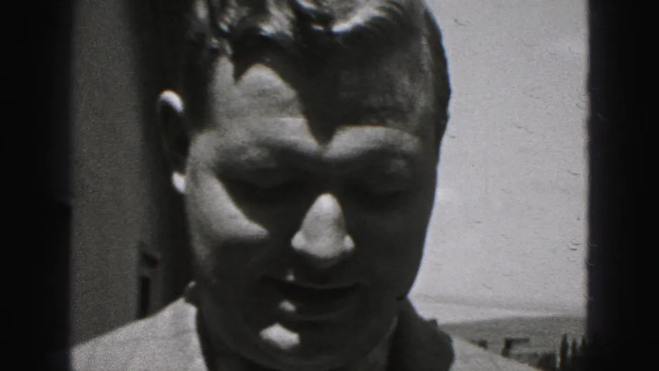 Close-up of a man facing the camera in 1975 Harrisburg home-movie footage, with strong sunlight and dark doorway-like edges behind him.
