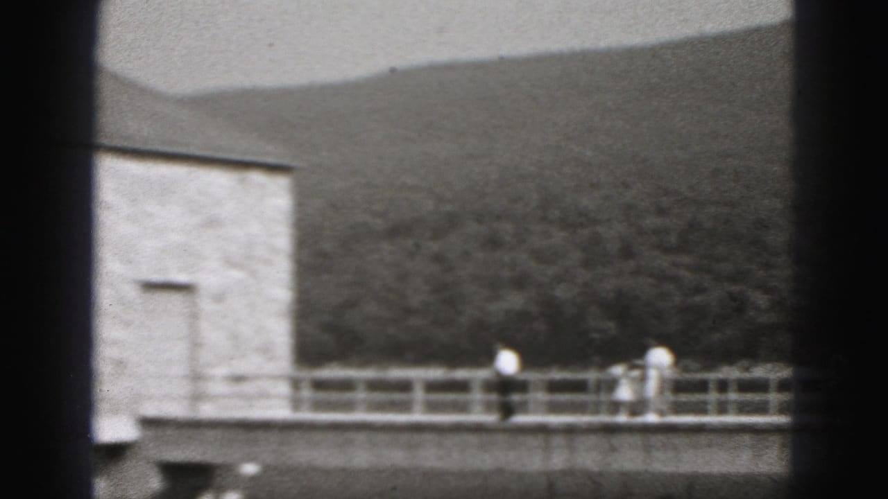 1975 Harrisburg footage shows distant pedestrians on a narrow waterside bridge near a square structure, with wooded hills beyond.