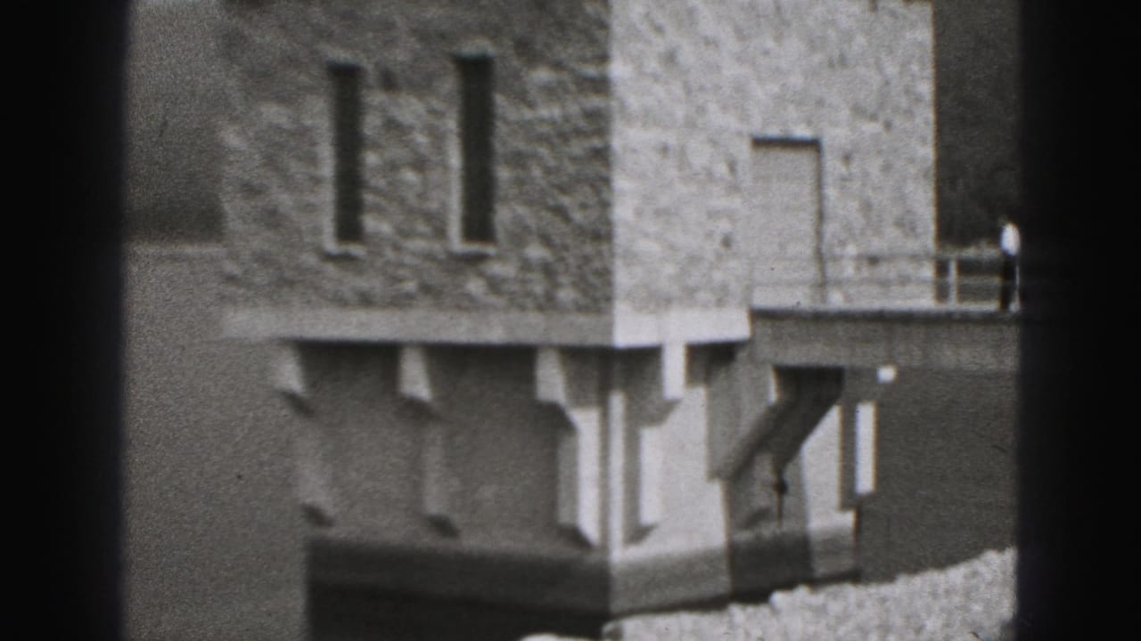 Stone building beside water with dark film borders and a gravel embankment in the foreground.