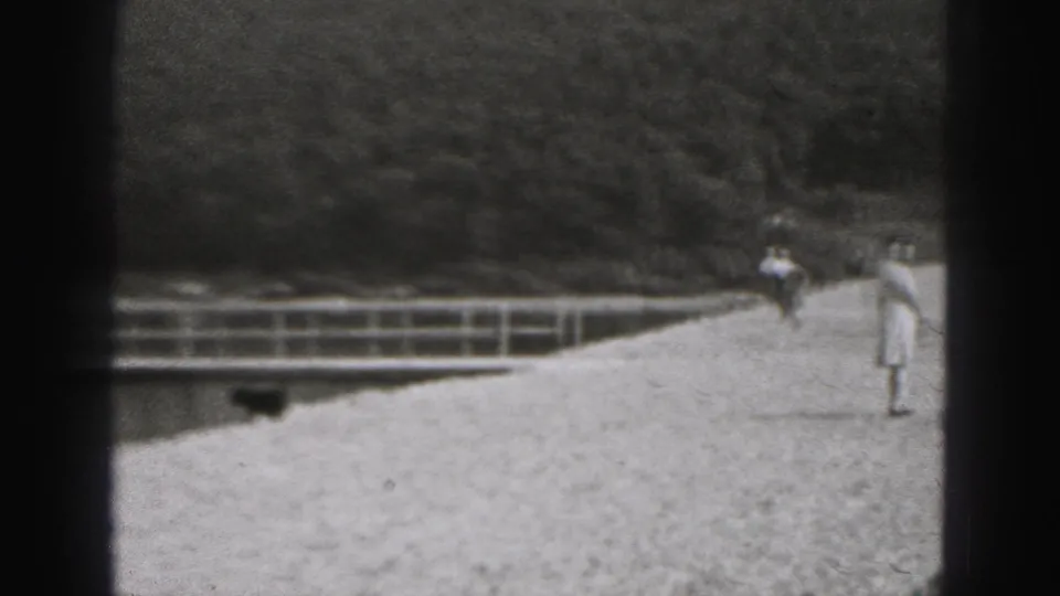 1975 Harrisburg home-movie frame with distant pedestrians on a gravel waterside path near a metal railing and wooded bank.