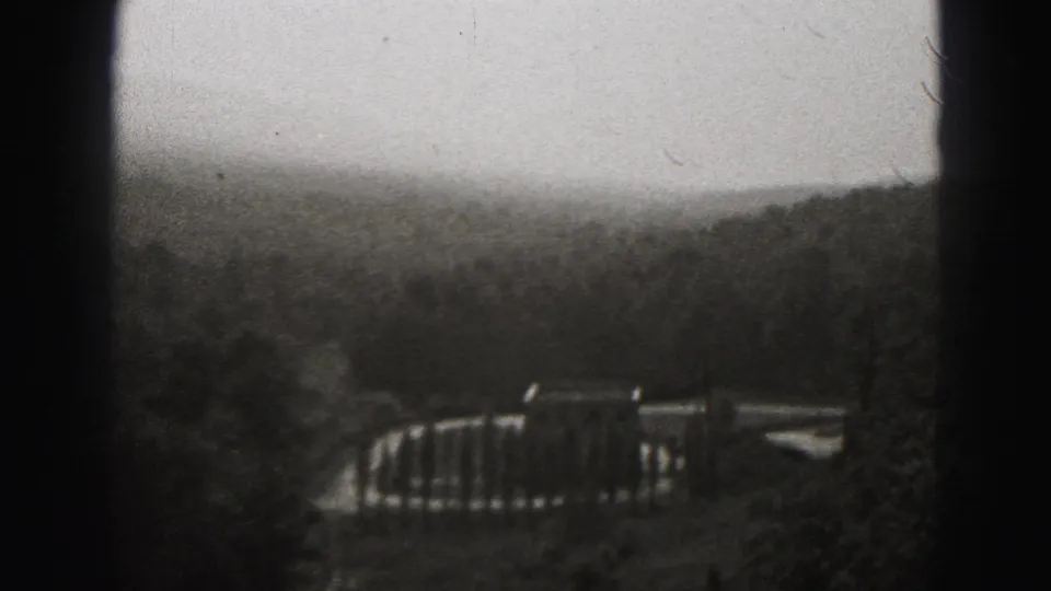 Final 1975 Harrisburg frame looking across a snowy hillside toward a small bridge or railing, with dark wooded terrain in soft focus.