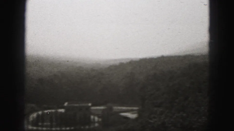 Harrisburg-area 1975 home-movie frame with distant structures among wooded terrain and dark 8mm gate edges.