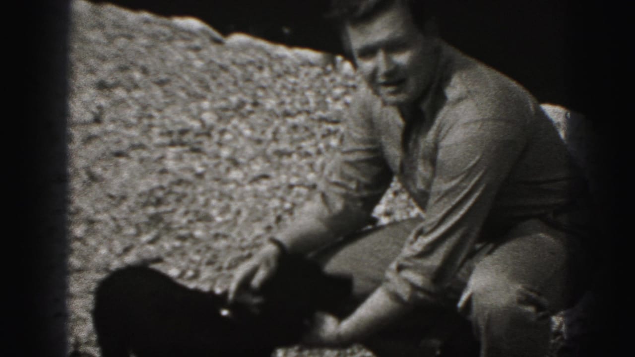 Adult man in a button-up shirt crouches beside a small black puppy outdoors in the 1975 Harrisburg 8mm footage.