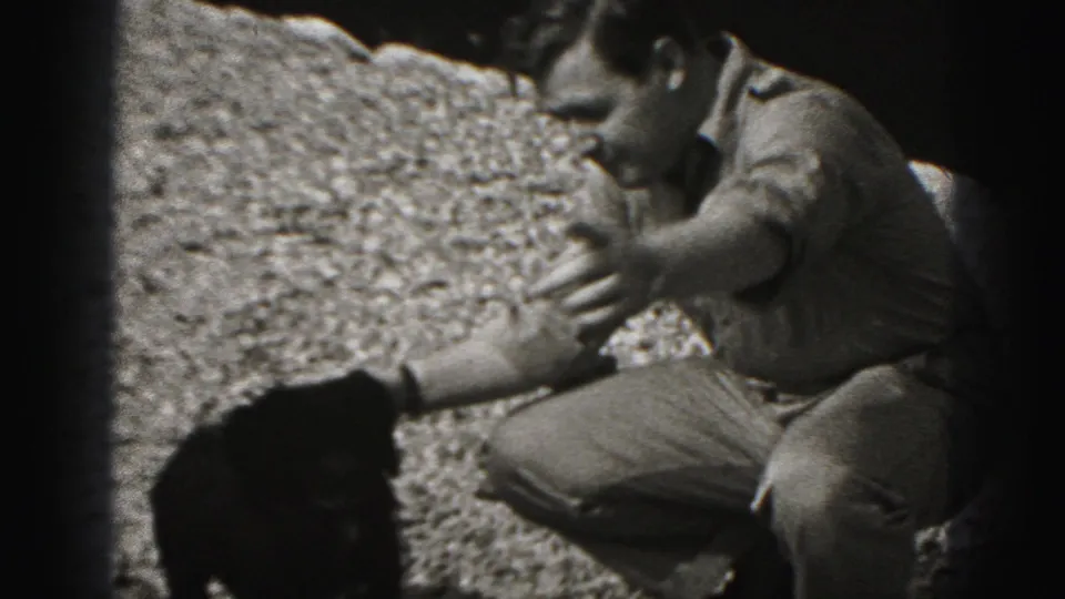 In the 1975 Harrisburg home movie, a crouching adult man reaches toward the small black puppy on gravel.