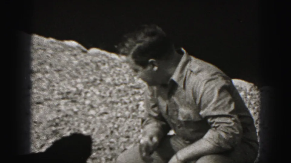 Adult man looks down toward the small black puppy as he crouches on gravel in the 1975 Harrisburg 8mm scene.