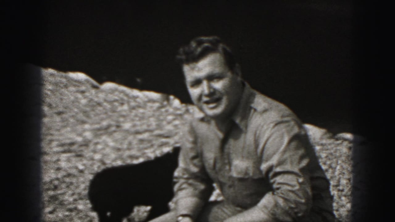 In 1975 Harrisburg home-movie footage, a crouching man faces the camera while a small black puppy stands near him on gravel.