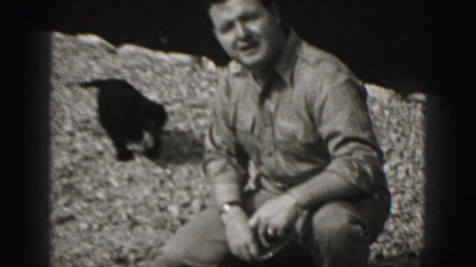 In Harrisburg, Pennsylvania home-movie footage from 1975, an adult man crouches on gravel while a small dark dog stands nearby.