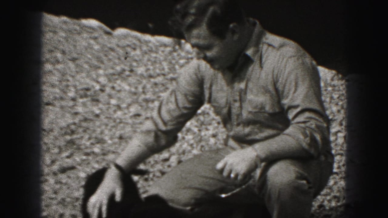 The man remains crouched on gravel in the 1975 Harrisburg home movie, lowering one hand toward the small dark dog.