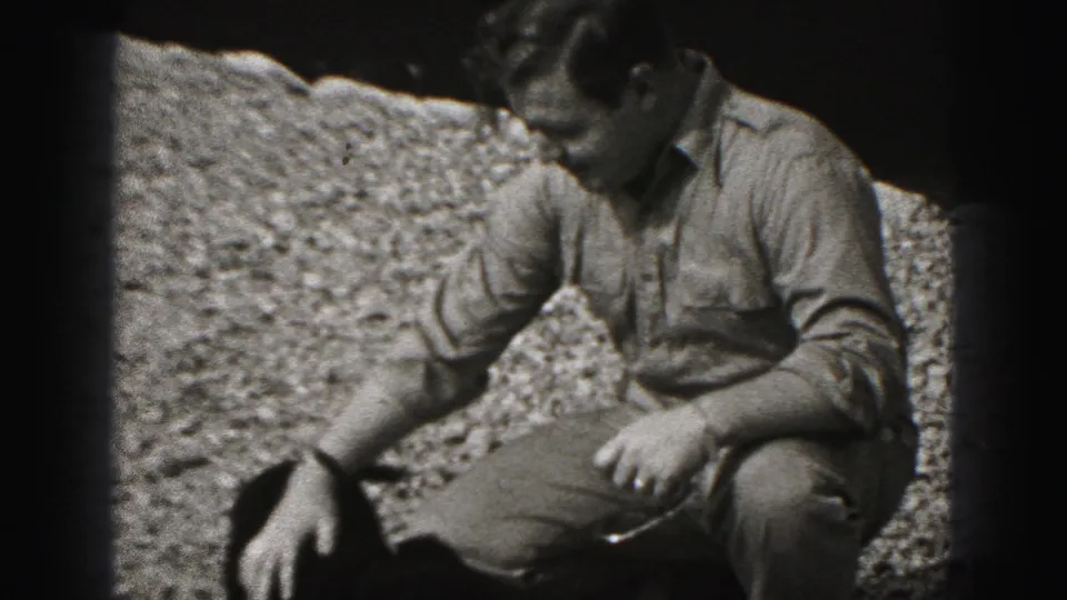 The man remains crouched on gravel in the 1975 Harrisburg home movie, lowering one hand toward the small dark dog.