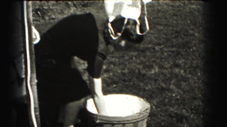 Woman bends toward a laundry basket beneath a clothesline in a Harrisburg yard, with hanging clothes and grass visible in the 1975 footage.
