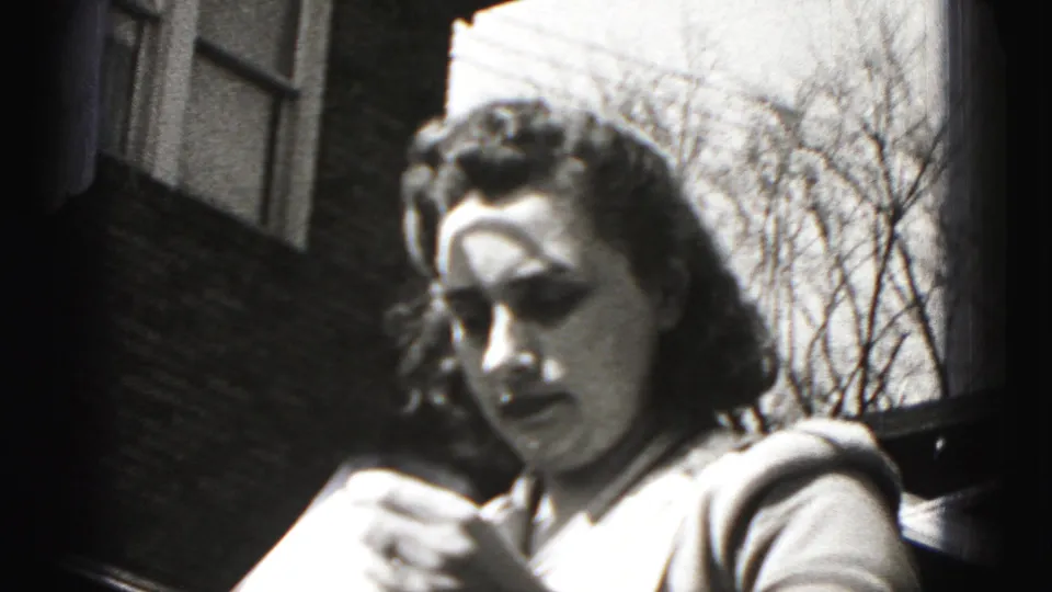 Young woman in Harrisburg, Pennsylvania, outdoors by a brick building, glancing down at paper in 1975 8mm footage.