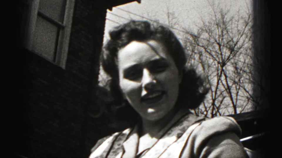 Woman in a cardigan faces the camera outside a brick building in Harrisburg, Pennsylvania, in 1975 home-movie footage.