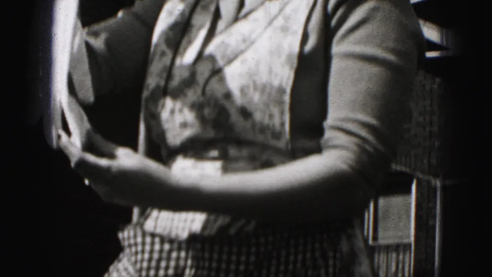 Woman in a patterned dress and cardigan holds paper outside a brick building in supported 1975 Harrisburg home-movie footage.