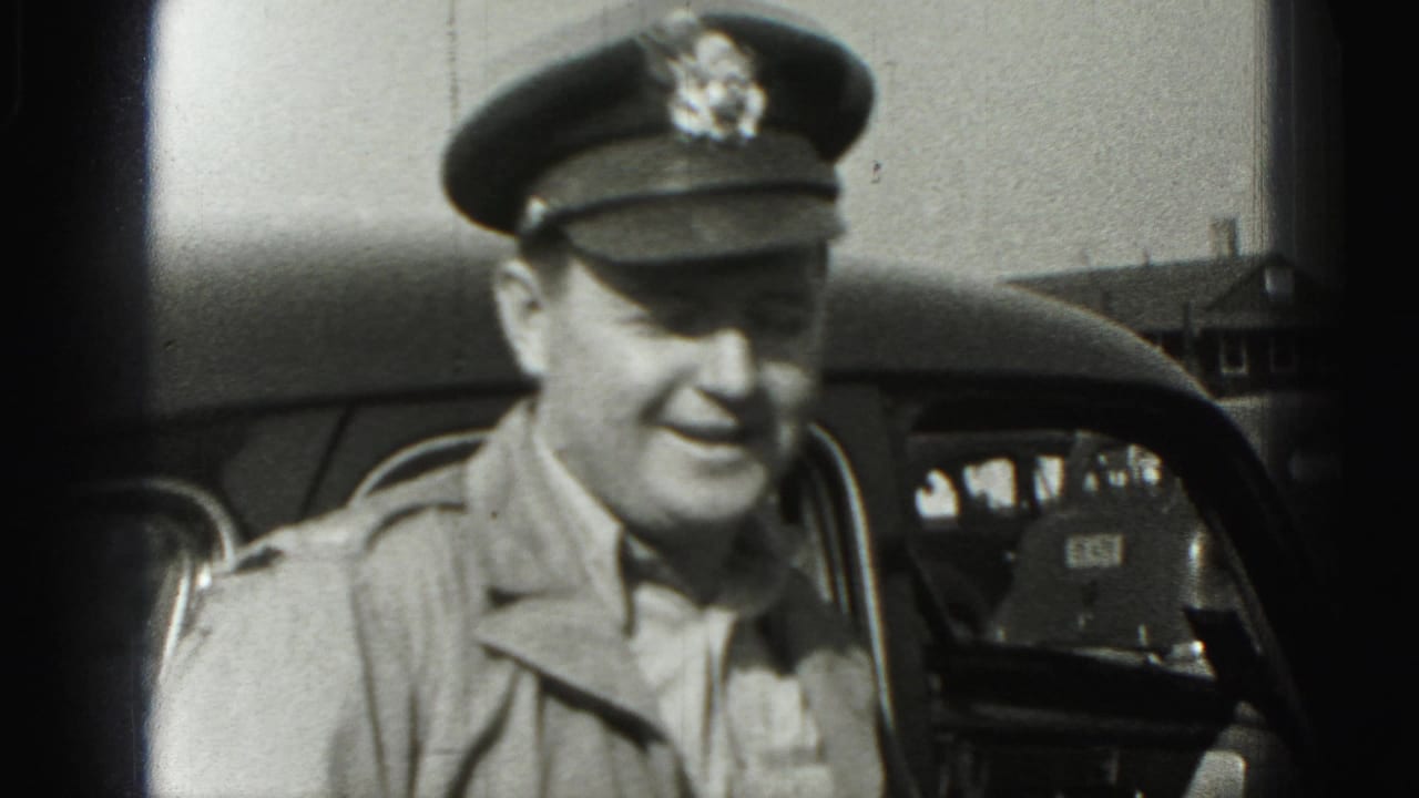 The uniformed man smiles toward the camera beside a vintage car, with another vehicle and a small building visible in the background.