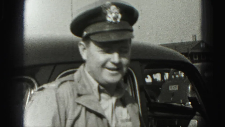 The uniformed man smiles toward the camera beside a vintage car, with another vehicle and a small building visible in the background.