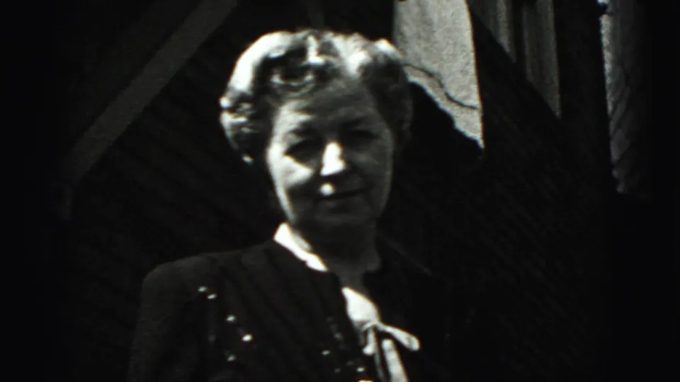 A woman in a dark dress with light trim stands outdoors in black-and-white 8mm footage, likely part of the same Harrisburg farewell scene.