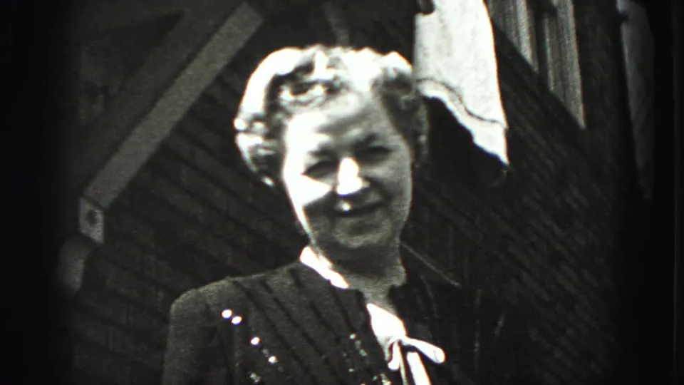 The woman smiles toward the camera near a building exterior, closing the Harrisburg home-movie farewell sequence in bright sunlight.