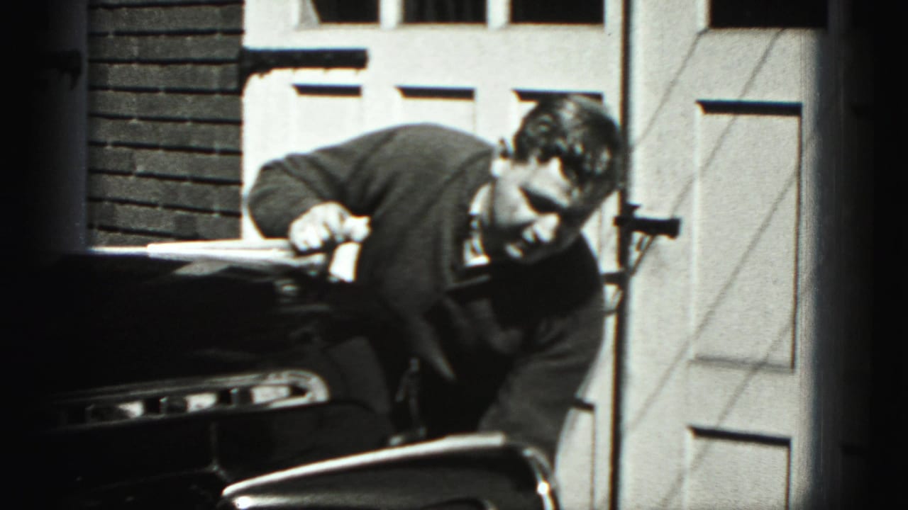In Harrisburg, Pennsylvania home-movie footage from 1975, a man leans over the front of a dark classic car in a driveway.