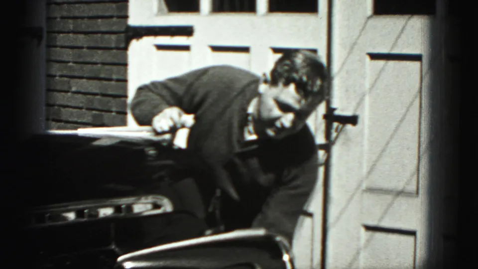 In Harrisburg, Pennsylvania home-movie footage from 1975, a man leans over the front of a dark classic car in a driveway.
