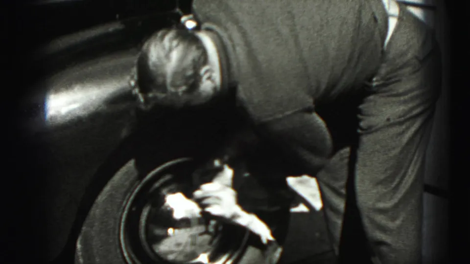 The 1975 home-movie frame shows the man bent close to the front wheel of a dark classic car, working with a cloth.