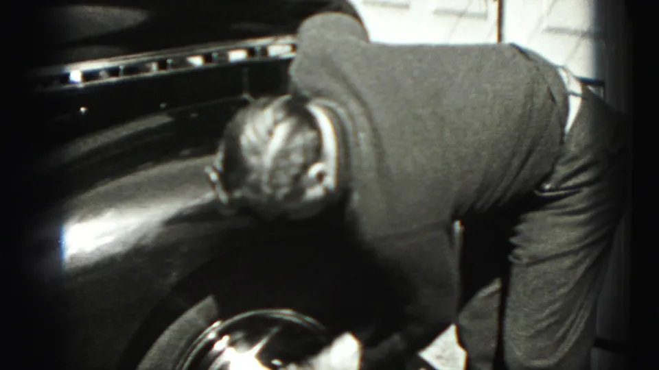 A man leans over the front fender and wheel of a classic car in Harrisburg, Pennsylvania, in 1975 home-movie footage.