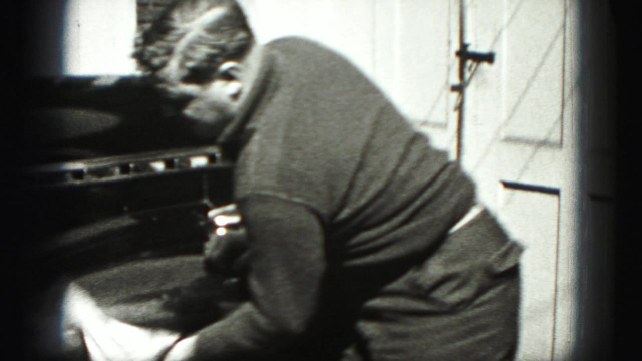 In Harrisburg, Pennsylvania home-movie footage, a man bends toward a car while washing or wiping it beside a garage door.