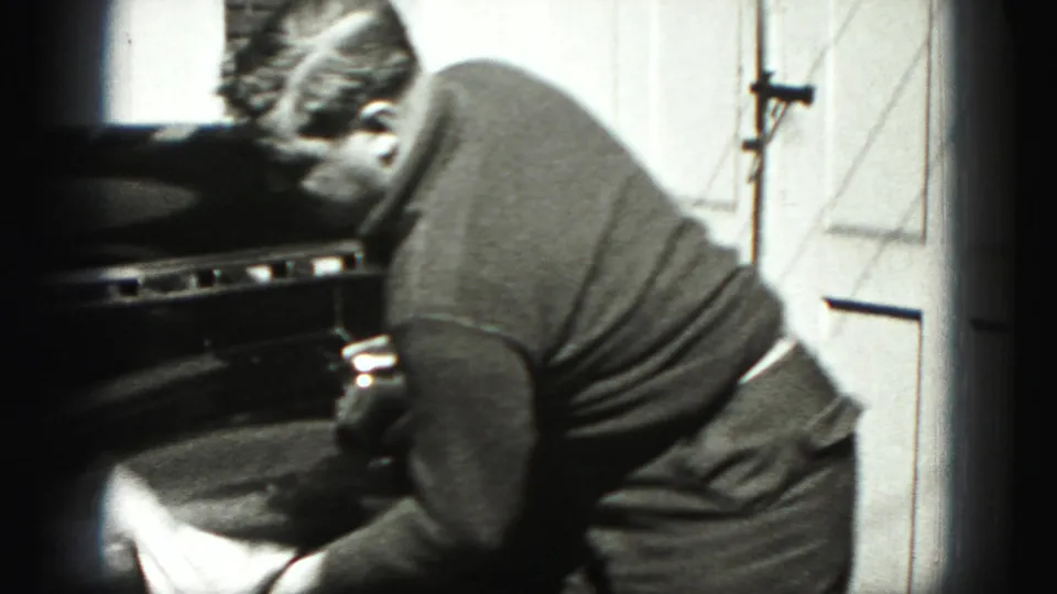 In Harrisburg, Pennsylvania home-movie footage, a man bends toward a car while washing or wiping it beside a garage door.