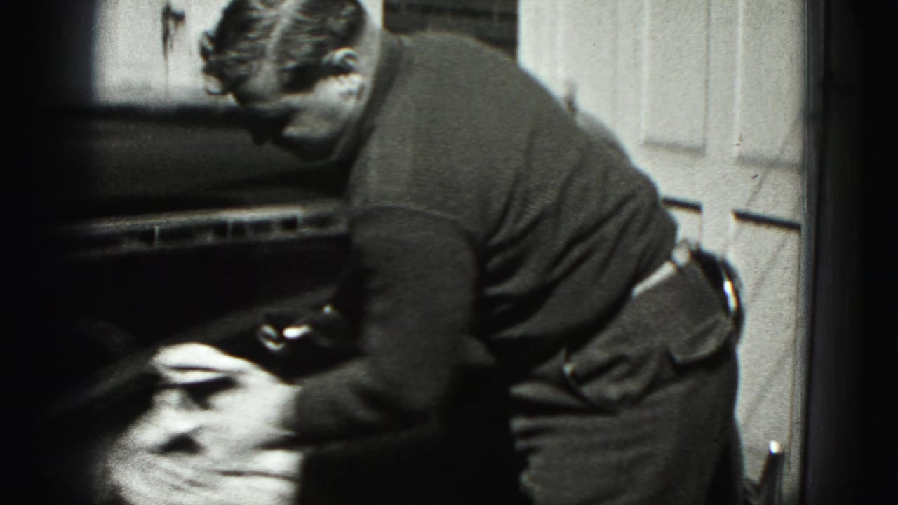 The man continues wiping the car body with a cloth in a residential driveway, filmed in grainy 8mm style.