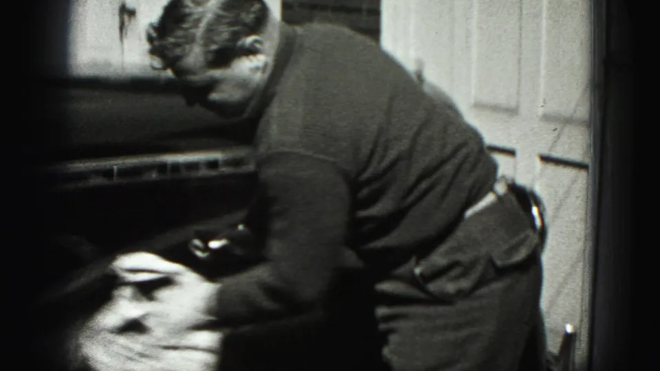 The man continues wiping the car body with a cloth in a residential driveway, filmed in grainy 8mm style.