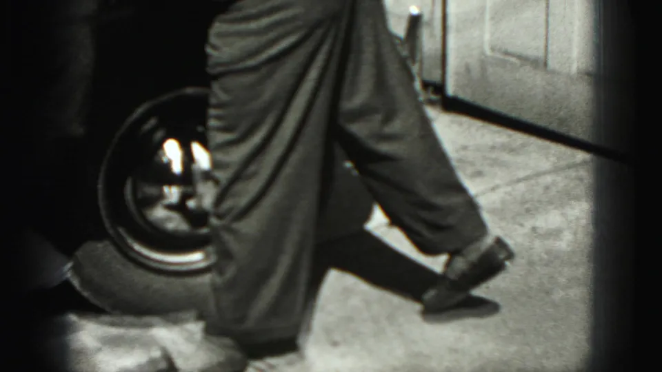 The man stands next to the car wheel and driveway area, with the garage door and vehicle body visible in the 1975 home-movie frame.