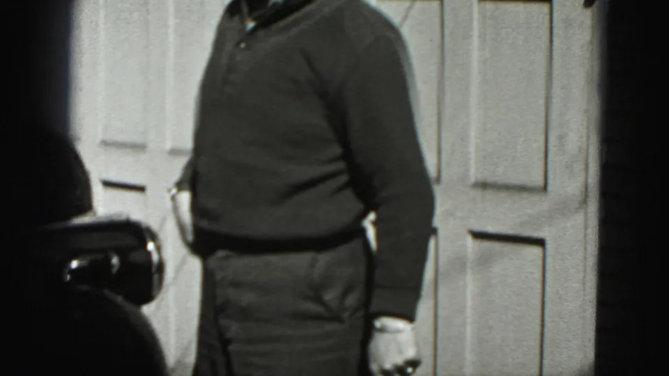 Adult man in a dark sweater stands near a classic car outside a garage in Harrisburg, Pennsylvania, 1975 home-movie footage.