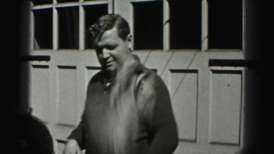 Adult man faces the camera near a paneled garage door during 1975 Harrisburg home-movie footage of car waxing.