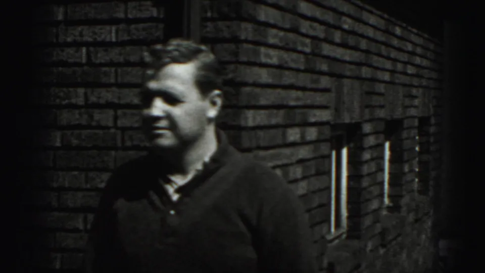 In supplied 1975 Harrisburg home-movie footage, a man in a dark sweater stands outdoors against a brick wall.