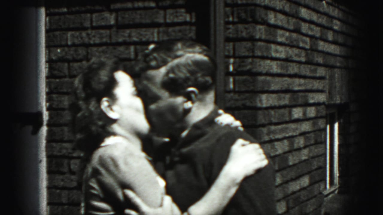 In Harrisburg 1975 home-movie footage, a young couple kisses in bright daylight against a brick exterior wall.