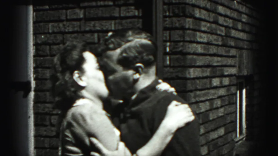 In Harrisburg 1975 home-movie footage, a young couple kisses in bright daylight against a brick exterior wall.