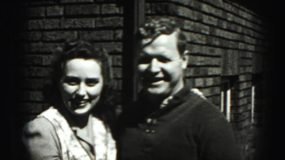 The couple remains close together, smiling in the sun as the 8mm camera frames them against the brick exterior.