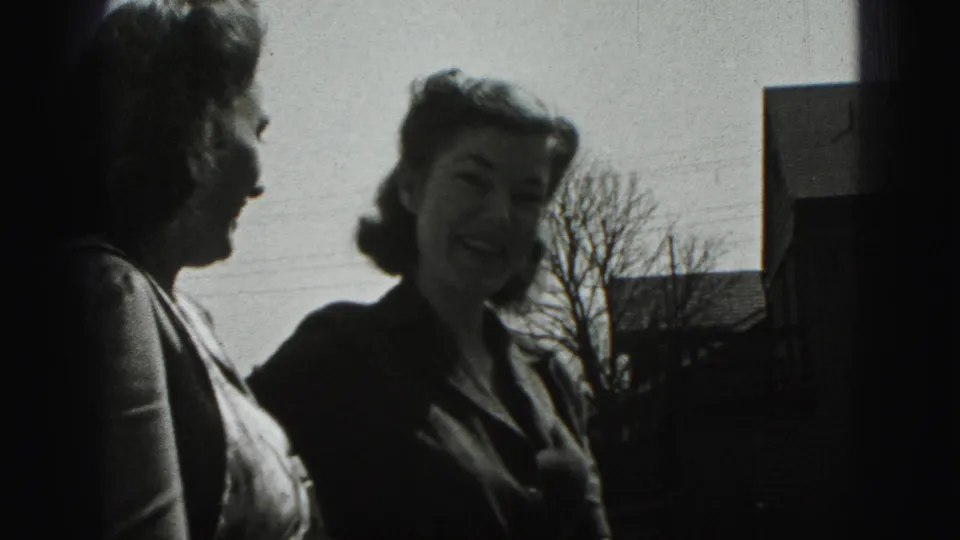 Two adult women in 1975 Harrisburg stand outside laughing, with bare trees and house roofs behind them.