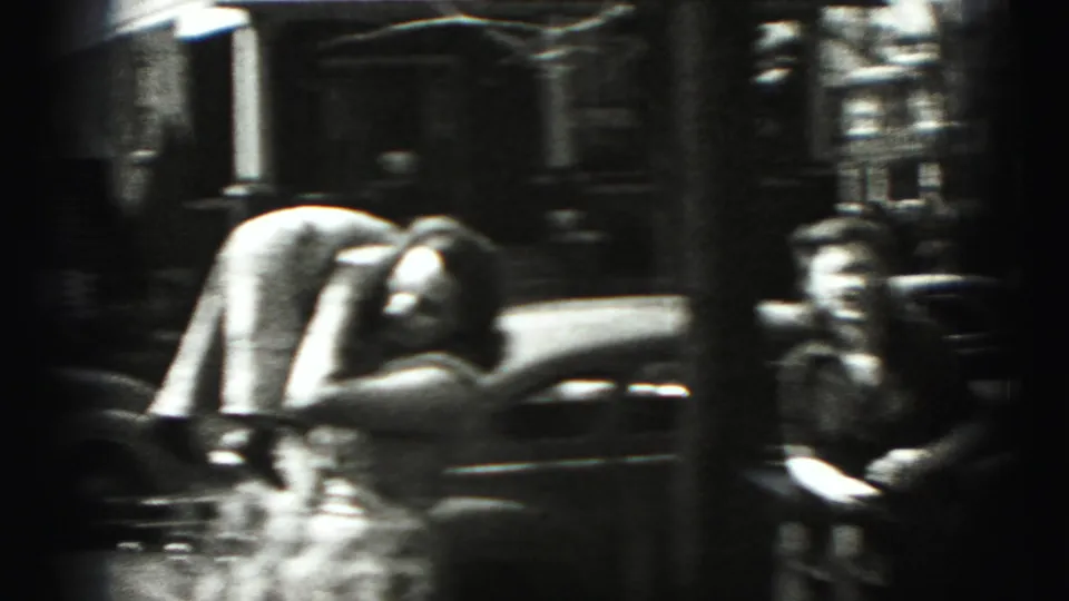 1975 Harrisburg home-movie frame with two figures beside a parked car on a quiet residential street.