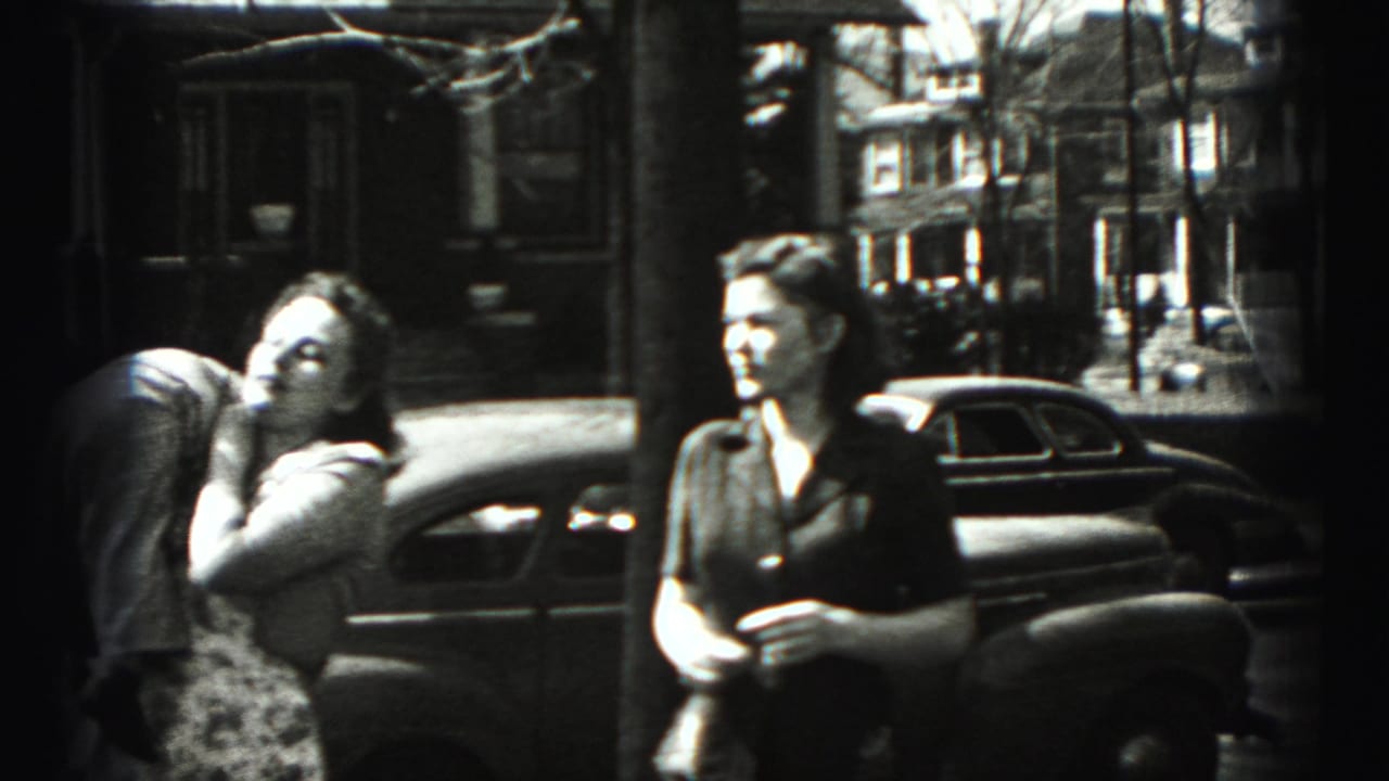 On a Harrisburg residential street, a woman in a patterned dress bends toward a smaller figure as another woman watches.
