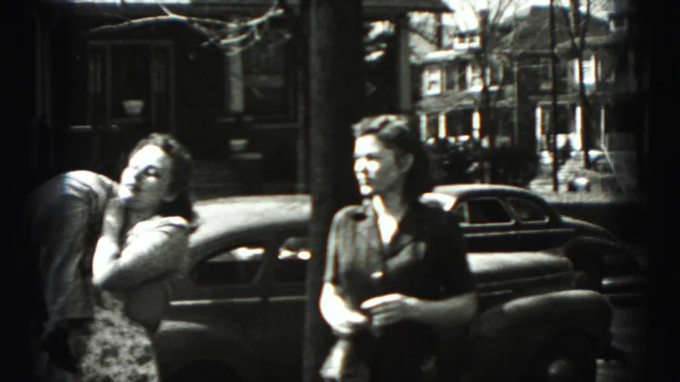 On a Harrisburg residential street, a woman in a patterned dress bends toward a smaller figure as another woman watches.