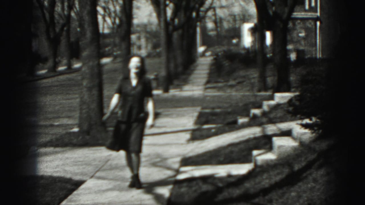 Woman walking toward the camera on a suburban sidewalk beside lawns and trees.
