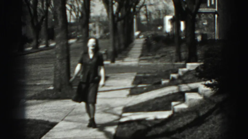 Adult woman walks along a tree-lined Harrisburg sidewalk in 1975 home-movie footage, with lawns and porch steps nearby.