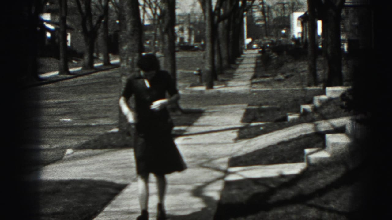 Woman in a dark dress stands or walks on a sidewalk, looking down at her clothing.