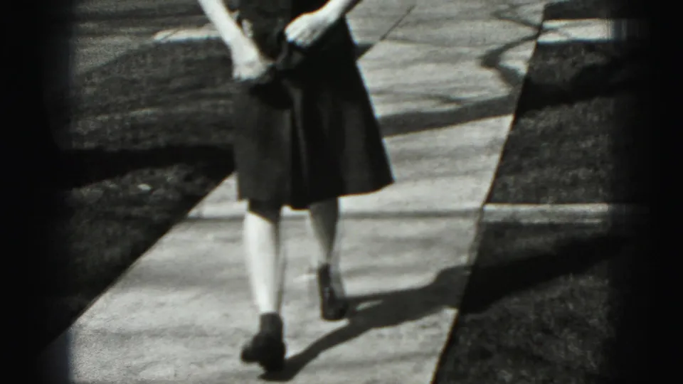 Closer 1975 home-movie frame of an adult woman in a dark dress walking along a suburban Harrisburg sidewalk.