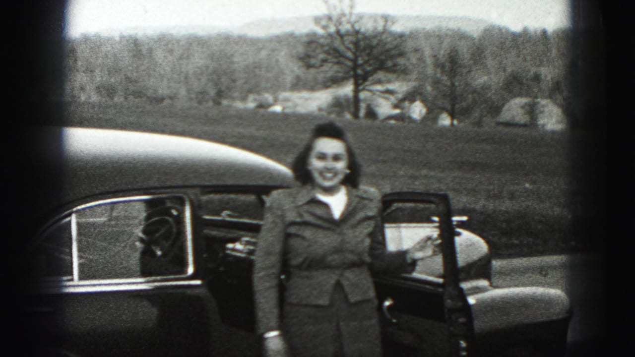 A smiling woman stands beside an open sedan door in Harrisburg, Pennsylvania, with open ground and trees in the background.