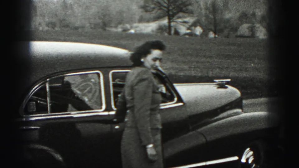 The woman walks past the side of the sedan in the Harrisburg home-movie scene, with fields and trees beyond the road.
