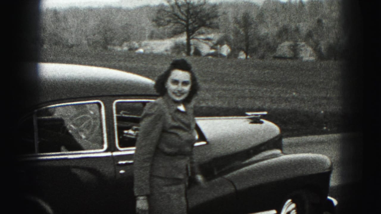 Woman in a suit standing beside a dark classic sedan on a rural roadside.
