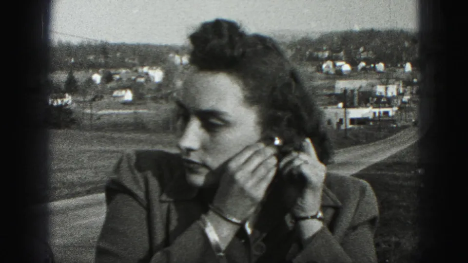 Close 8mm frame of the woman adjusting an earring, with a rural roadside and distant buildings behind her.